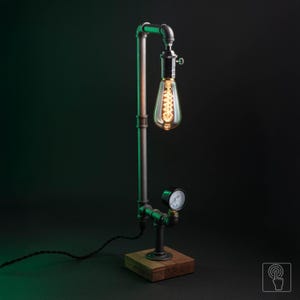 Może przedstawiać: Lampa stołowa wykonana z ciemnoszarych metalowych rur hydraulicznych i złączek, zamontowana na kwadratowej drewnianej podstawie. Lampa posiada jedną skierowaną w dół żarówkę typu Edison oraz mały okrągły manometr przymocowany do rury.