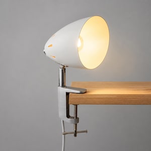May include: A white metal clamp lamp with a rounded shade featuring small triangular cutouts. The lamp is attached to a wooden surface by a silver metal screw clamp mechanism and emits a warm, bright light from the interior bulb.