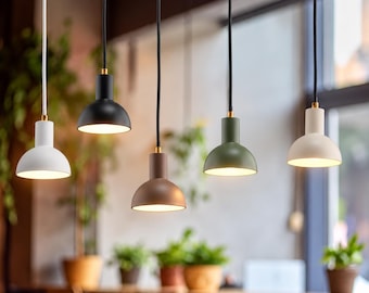 May include: Five pendant lights with dome-shaped shades in white, black, brown, olive green, and cream. Each lamp features a cylindrical top section, a gold-toned connector, and a cord. The lights are illuminated, casting a warm glow downwards.