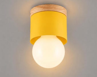 May include: A flush mount ceiling light fixture featuring a cylindrical yellow metal shade attached to a light-toned wooden base. The fixture includes a large, spherical, frosted white glass light bulb that emits a warm glow.