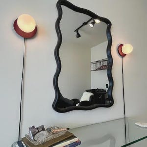 Op de afbeelding: Een spiegel met een zwarte, golvende lijst hangt aan een muur tussen twee wandlampen. Elke lamp heeft een ronde, donkerrode voet en een bolvormige witte glazen kap. Een zwart snoer loopt van elke lamp naar beneden naar het oppervlak.
