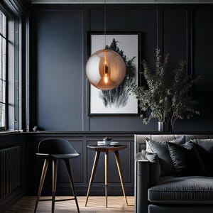 A stylish living room featuring dark blue walls, a black chair, and a grey sofa. A large, spherical, amber-toned pendant light hangs from the ceiling. A small round table with wooden legs and a monochrome print are also present.