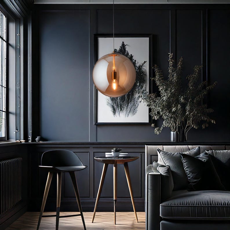 A stylish living room featuring dark blue walls, a black chair, and a grey sofa. A large, spherical, amber-toned pendant light hangs from the ceiling. A small round table with wooden legs and a monochrome print are also present.