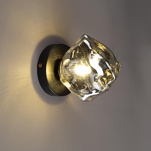 May include: A modern wall sconce featuring a textured, irregularly shaped glass shade. The shade is a translucent amber color, and it is mounted on a circular black metal base. The light is turned on, illuminating the glass.