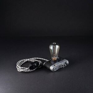 May include: A vintage-style camera repurposed as a table lamp, featuring a silver and black camera body with a clear Edison-style light bulb extending from the top. A coiled grey and white fabric-covered power cord with a black switch is attached.