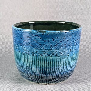 May include: A ceramic bowl with a textured, blue glaze. The bowl features a gradient of blues, transitioning from a dark teal interior to a lighter turquoise at the base. The design includes vertical and geometric patterns.