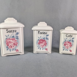 Puede incluir: Tres botes de cerámica blanca con diseños de rosas rosas y detalles azules. Los botes están etiquetados como "Sucre", "Farine" y "Cafe".