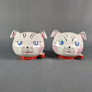 Puede incluir: Dos figuritas de cerámica blanca en forma de cabeza de gato con ojos negros y collares rojo y blanco. Las cabezas de gato están mirando hacia adelante y tienen una expresión caprichosa.