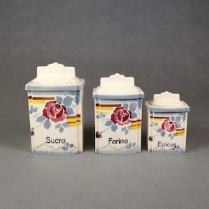Puede incluir: Juego de tres botes de cocina de cerámica con tapas. Cada bote es blanco con un diseño floral con rosas rojas y hojas azules. Los botes están etiquetados como "Sucre", "Farine" y "Epices".