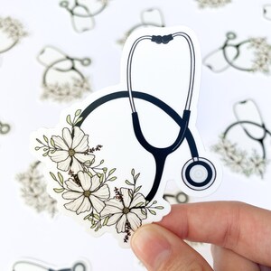 May include: A white sticker with a black stethoscope and white flowers. The stethoscope has a black tube and a silver listening piece. The flowers are arranged in a circle around the stethoscope.