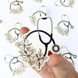 May include: A white sticker with a black stethoscope and white flowers. The stethoscope is in the shape of a circle with the earpieces at the top. The flowers are arranged around the stethoscope.