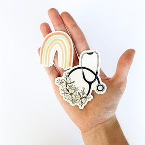 May include: Three stickers on a hand. One sticker is a rainbow with pastel colors. Another sticker is a stethoscope with white flowers around it. The third sticker is a stethoscope with a black outline.