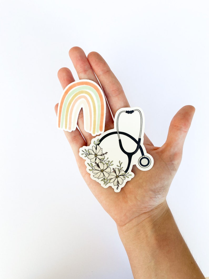 May include: Three stickers on a hand. One sticker is a rainbow with pastel colors. Another sticker is a stethoscope with white flowers around it. The third sticker is a stethoscope with a black outline.