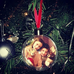 May include: Clear glass Christmas bauble featuring a photo of two children holding stuffed reindeer. The bauble has a red ribbon.