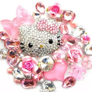 Puede incluir: Una colecci&oacute;n de adornos brillantes, que incluye una cara de Hello Kitty cubierta de pedrer&iacute;a, rosas rosas y varias gemas facetadas. La disposici&oacute;n presenta una mezcla de elementos rosas, transparentes y plateados, creando una est&eacute;tica glamurosa y decorativa.