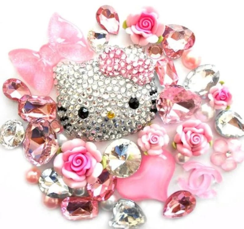 Puede incluir: Una colecci&oacute;n de adornos brillantes, que incluye una cara de Hello Kitty cubierta de pedrer&iacute;a, rosas rosas y varias gemas facetadas. La disposici&oacute;n presenta una mezcla de elementos rosas, transparentes y plateados, creando una est&eacute;tica glamurosa y decorativa.