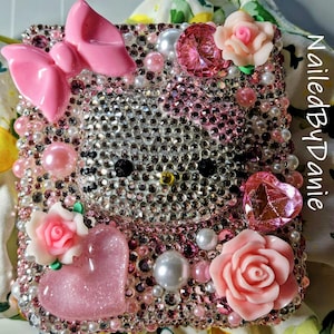 Puede incluir: Una funda de tel&eacute;fono adornada con un dise&ntilde;o de Hello Kitty. La funda est&aacute; cubierta de pedrer&iacute;a, perlas y adornos rosas, incluyendo un lazo, corazones y rosas. El texto "Nailed By Danie" es visible en el lateral.