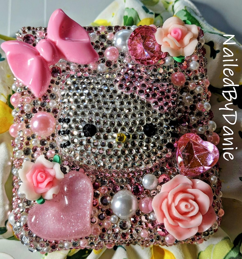 Puede incluir: Una funda de tel&eacute;fono adornada con un dise&ntilde;o de Hello Kitty. La funda est&aacute; cubierta de pedrer&iacute;a, perlas y adornos rosas, incluyendo un lazo, corazones y rosas. El texto "Nailed By Danie" es visible en el lateral.