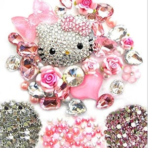 Puede incluir: Un surtido de adornos decorativos con una cara de gato cubierta de brillantes pedrer&iacute;a, rosas rosas y varias formas de cristal rosa y transparente. El arreglo incluye un lazo rosa, un coraz&oacute;n y grupos de cuentas rosas y plateadas.