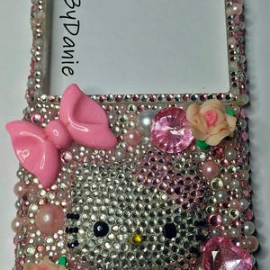 Puede incluir: Una funda de tel&eacute;fono adornada con un dise&ntilde;o de Hello Kitty. La funda est&aacute; cubierta de pedrer&iacute;a, perlas y detalles en rosa, incluyendo un lazo, un coraz&oacute;n y rosas. El texto "NailedByDanie" es visible en el lateral.