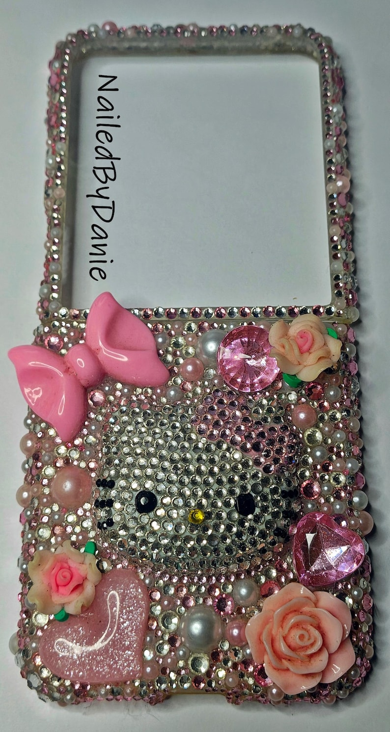 Puede incluir: Una funda de tel&eacute;fono adornada con un dise&ntilde;o de Hello Kitty. La funda est&aacute; cubierta de pedrer&iacute;a, perlas y detalles en rosa, incluyendo un lazo, un coraz&oacute;n y rosas. El texto "NailedByDanie" es visible en el lateral.