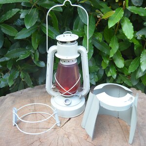 May include: White metal lantern with a red glass globe and a white metal stand. The lantern has a handle and a clear glass chimney. The stand has three legs and is designed to hold the lantern.