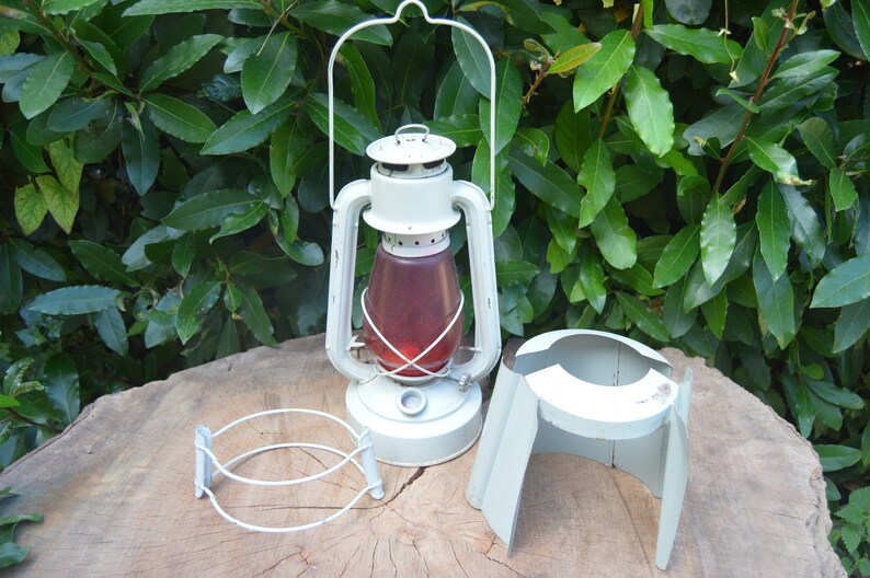 May include: White metal lantern with a red glass globe and a white metal stand. The lantern has a handle and a clear glass chimney. The stand has three legs and is designed to hold the lantern.