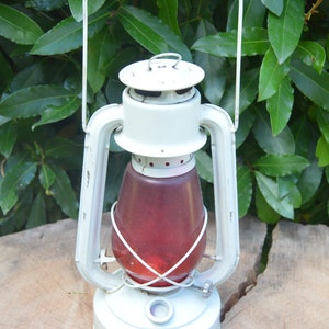 May include: A white metal lantern with a red glass globe and a wire cage. The lantern has a handle on top and a base with a small knob.