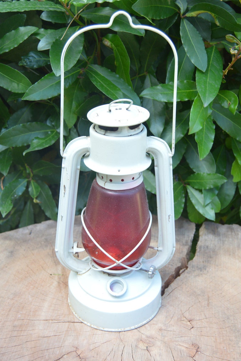 May include: A white metal lantern with a red glass globe and a wire cage. The lantern has a handle on top and a base with a small knob.