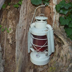 May include: A white metal lantern with a red glass panel hangs from a tree branch. The lantern has a wire handle and a metal frame.