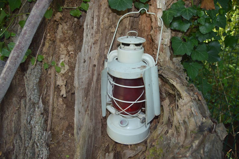 May include: A white metal lantern with a red glass panel hangs from a tree branch. The lantern has a wire handle and a metal frame.