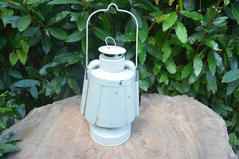 May include: A white metal lantern with a handle and a round top. The lantern is sitting on a wooden surface.