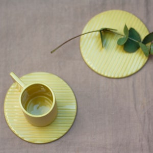 May include: A yellow ceramic cup and saucer with a ribbed design. The cup is empty and the saucer is on a light brown surface.