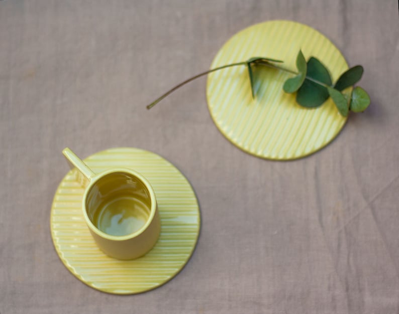 May include: A yellow ceramic cup and saucer with a ribbed design. The cup is empty and the saucer is on a light brown surface.