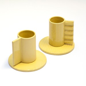 May include: Two yellow ceramic coffee cups with unique handles and matching saucers. The cups and saucers have a textured surface.