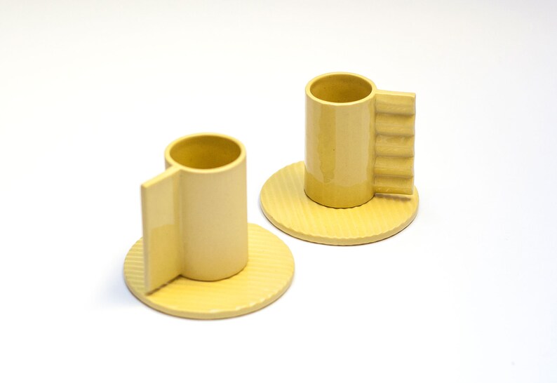 May include: Two yellow ceramic coffee cups with unique handles and matching saucers. The cups and saucers have a textured surface.