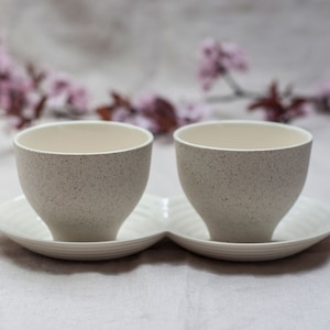 Könnte beinhalten: Zwei weiße Keramik-Teetassen mit gesprenkelter Textur, die auf einer weißen Untertasse mit gewelltem Rand stehen.