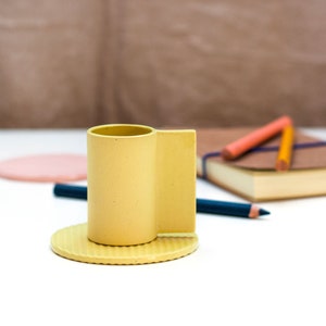 May include: A yellow ceramic cup with a rounded base and a rectangular handle. The cup is sitting on a matching yellow saucer.