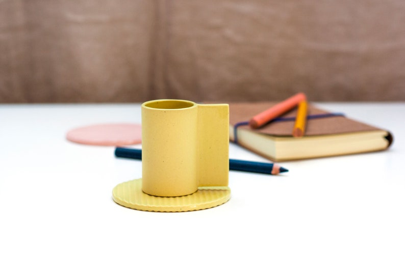 May include: A yellow ceramic cup with a rounded base and a rectangular handle. The cup is sitting on a matching yellow saucer.