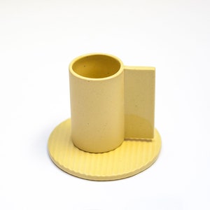 May include: A yellow ceramic cup and saucer set. The cup has a unique handle that extends from the side. The saucer has a textured surface.
