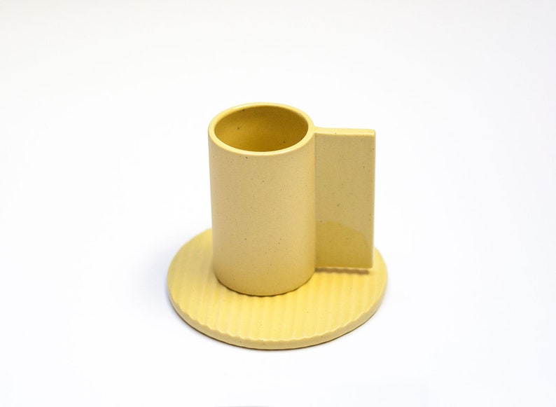 May include: A yellow ceramic cup and saucer set. The cup has a unique handle that extends from the side. The saucer has a textured surface.