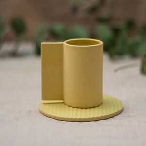 May include: A yellow ceramic cup with a unique handle and a matching saucer. The saucer has a ridged design.
