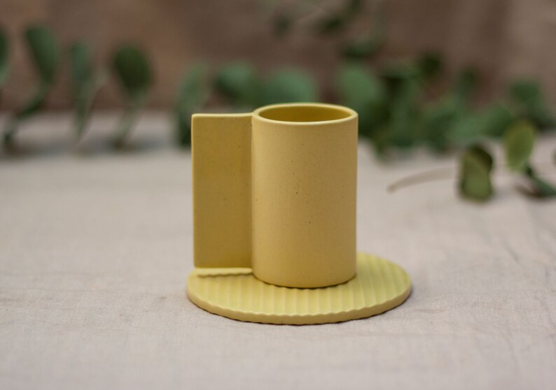 May include: A yellow ceramic cup with a unique handle and a matching saucer. The saucer has a ridged design.