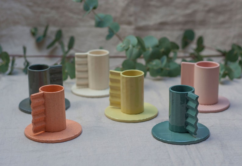 May include: Set of six ceramic cups with unique, textured handles. The cups are in various colors, including pink, green, yellow, and orange. Each cup has a matching saucer.