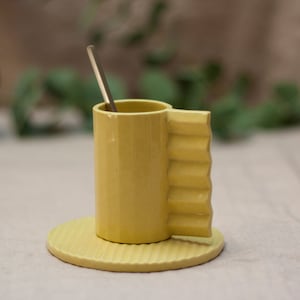 May include: A yellow ceramic mug with a fluted handle and a matching saucer. The mug has a textured surface and is sitting on a light beige surface.