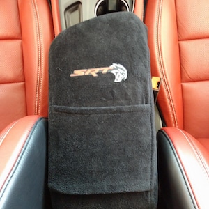 May include: A black fabric center console cover featuring the embroidered SRT logo and a cat head emblem. The cover has a horizontal pocket across the middle and is positioned between two red leather car seats with visible stitching details.
