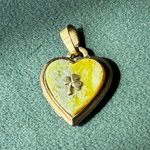 May include: A gold-colored heart-shaped pendant with a shamrock design. The pendant has a yellow and white marbled center and a small gold shamrock charm. The pendant is on a green textured surface.