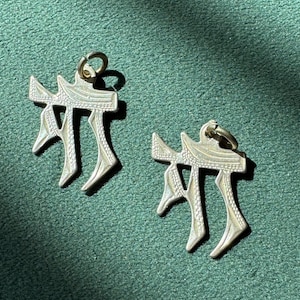 May include: Two silver pendants with a distinctive, stylized design. Each pendant has a loop at the top for hanging and a unique shape with three downward-pointing elements. The pendants are displayed against a green background.