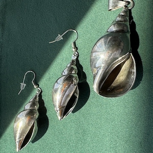 Könnte beinhalten: Ein Schmuckset bestehend aus zwei silberfarbenen, muschelförmigen Ohrringen mit Hakenverschluss und einem passenden, größeren muschelförmigen Anhänger mit einer silberfarbenen Öse, alle mit einer polierten, reflektierenden Metalloberfläche.