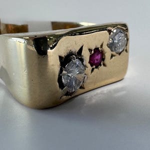 Può includere: Un anello in oro con una faccia rettangolare e un'incastonatura a forma di stella. L'anello è decorato con due pietre trasparenti a taglio rotondo sui lati esterni e una singola pietra rossa più piccola a taglio rotondo al centro.